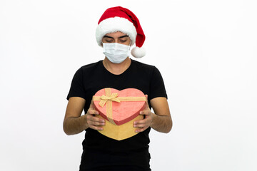 Young male wearing a medical face mask and a santa hat. Young man is holding a heart-shaped gift box in his hands. Isolated image and white background.