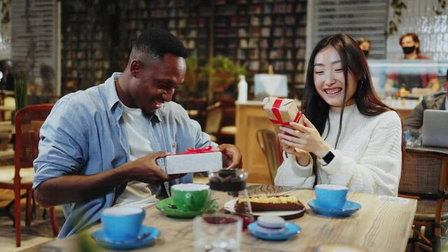 Multi-race young couple of attractive man and woman sitting in cafe drinking coffee exchanging gifts for holiday Christmas party together. Public place. People.