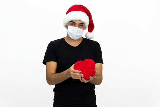 Young male wearing a medical face mask and a santa hat. Young man is holding a heart-shaped gift box in his hands. Isolated image and white background.