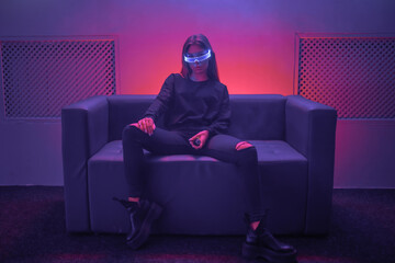 Cyberpunk woman sitting on sofa with neon glasses. The photo has the effect of shush, grain.
