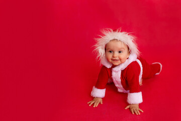 a small child of six months in a Santa costume crawls on a red isolated photophone and looks at the...