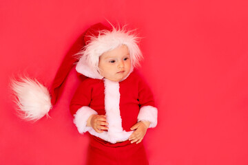 a small child of six months in a Santa costume lies on a red isolated photophone, a place for text, the concept of the new year and holiday