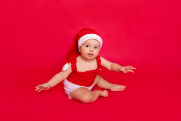 a small child of six months in a Santa hat sits on a red isolated photophone holding his hands to the sides, a place for text, the concept of the new year and holiday
