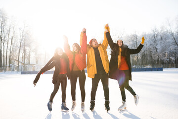 Joyful three girls and a guy in warm jackets stand on the rink raising their hands above their heads