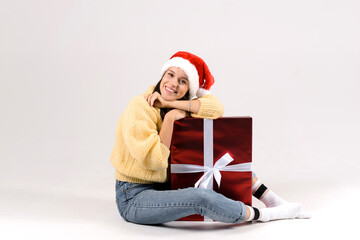 Young woman sitting near big red christmas gift box on the floor againt white studio background. Smiling happy girl in christmas style and red santa hat