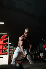 Fototapeta na wymiar A male African American crossfit trainer with a naked torso doing exercises with a hammer. Athlete trains hammer blows in the gym