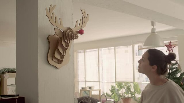 A lady with brown hair in a bun in a home wear is hanging a small red shiny Christmas ball on a carton decorative deer head in her appartment. Light, warm atmosphere. 4k high quality video footage.