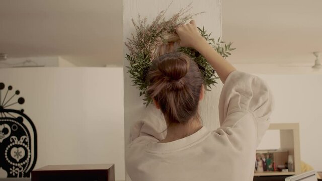 Caucasian girl with brown hair in a bun in a home wear is hanging a hand made winter festive wreath on the white column in her homet. Light, warm atmosphere. Back side view 4k high quality video.