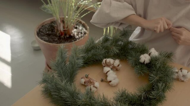 Dark hair european female florist no make up in a beige top sitting at the wooden table preparing cotton flowerto put on the Chrstmas wreath. Warm light atmosphere. Handled shot 4k high quality video