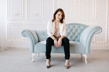 Attractive young woman in a business suit with white jacket, black pants and high heels sits on a blue classic sofa. White background with light
