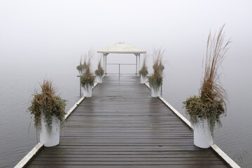 Romantic moody scenery with wooden pier with white gazebo by lake on calm misty day