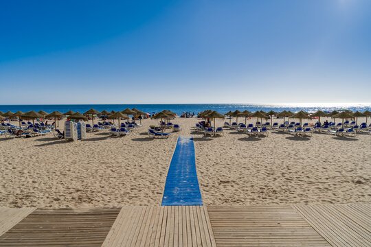 Playa con una pasarela sobre la arena que lleva a las sombrillas y el agua del mar un d&iacute;a soleado con cielo azul. Desde la playa de la Victoria, C&aacute;diz, Andaluc&iacute;a, Espa&ntilde;a.