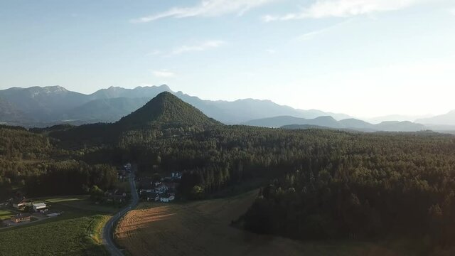 A drone capture of Katherinekogel, towering above the forest in Austria. The small hill resembles a pyramid in shape. In the back there are high Alpine chains. There are many farmlands in front
