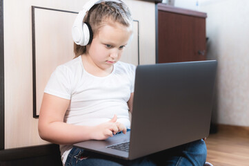 A smart little girl of seven years old in white headphones with a laptop in her hands is pushing on the floor in her room. The young generation on the Internet and IT technology