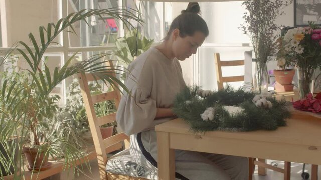 A beautiful brunette european florist girl no make up in a home wear is sitting at the wooden table putting together Chrstmas wreath. Warm light atmosphere. Handled shot 4k high quality video footage.