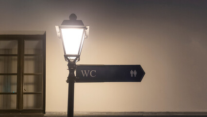 Street lamp with street sign with the inscription WC on empty wall background. Night time. Design concept.