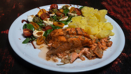 Boiled potatoes, fresh salad and roasted salmon on white plate. Dark wooden background. Cuisine concept.