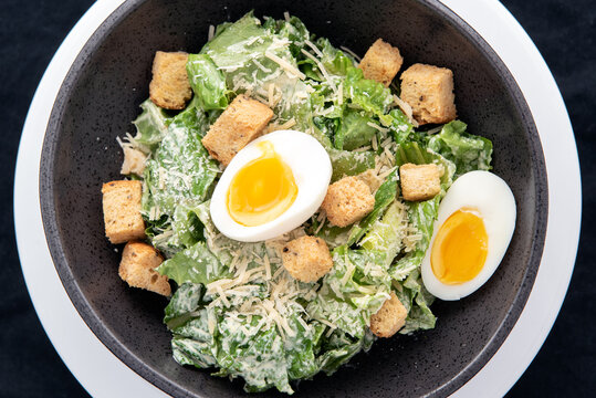 Overhead View Of Bowl Of Caesar Salad With Topped With Parmesan Cheese, Croutons, And Hard Boiled Egg.