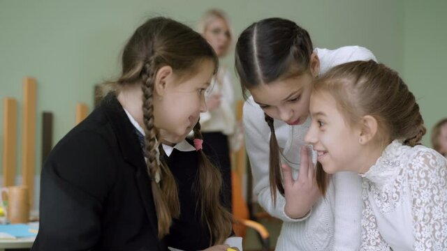 Close-up of four schoolgirls gossiping with blurred teacher talking at the background. Cute Caucasian girls sharing secrets and talking in classroom in public school during break. Friendship concept.