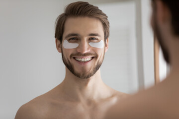 Fototapeta na wymiar Head shot close up mirror reflection happy millennial guy standing in bathroom with under eyes hydrogel patches, enjoying anti-wrinkle spa domestic morning skincare procedure, looking at camera.