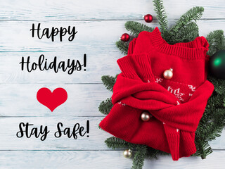 Red christmas winter sweater with green ornament on white wooden background. Happy holidays, stay safe text. Greeting card in times of covid pandemic.
