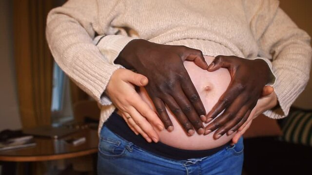 The black hands of the future father fold the fingers in the shape of a heart on the belly of a white-skinned woman expecting a baby. Challenges of an interracial marriage from society.