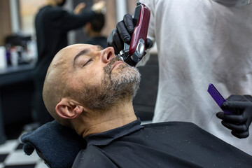 Close up picture of a barber's hands shaving bearded man with a trimmer in salon