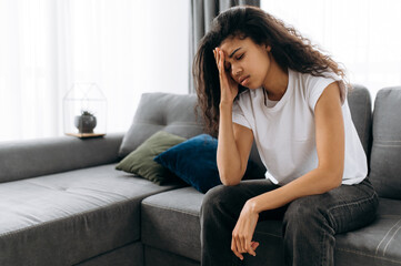 Sad african american young woman sit on couch at home feels unhappy because of headache, personal...