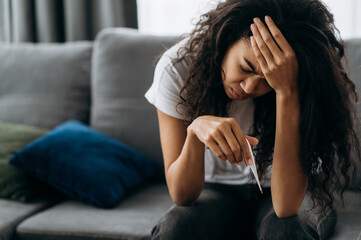 Unhappy sick african american girl sit on couch feel unhealthy, she looks at the thermometer in...