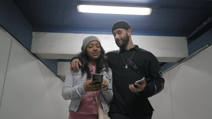 A young interracial couple walking in a subway corridor, looking forward to something on their smart phone. - Powered by Adobe