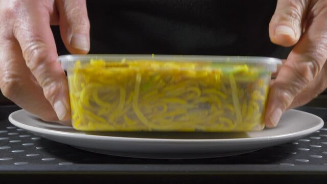 Closeup POV shot of a man removing the lid from a plastic takeout carton of hot Chinese noodles, then tipping the meal onto a plate.