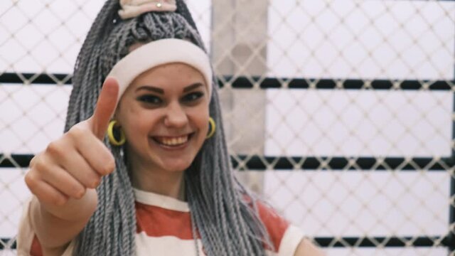 Portrait of excited young woman with dreadlocks or kanekalon pigtails looking at camera and smiling showing thumbs up wearing white headband and yellow earrings