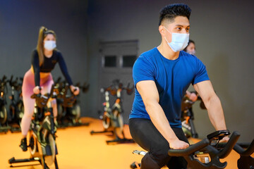 Young People Spinning in the fitness gym with protective face mask during coronavirus outbreak ....