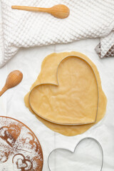 A heart-shaped cutting mold on rolled dough next to a towel and wooden spoons. Flat lay, vertical