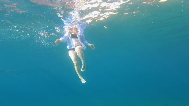 Young woman in two-piece swimsuit and white shirt swims in the sea, someone stealthily spies on her under water, shooting on action camera, bottom view