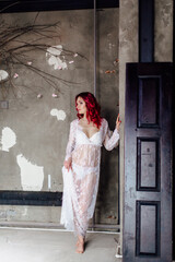 Attractive woman with pink hair in white light dress posing in a dark room with concrete floor and walls. Lady in gentle lace peignoir