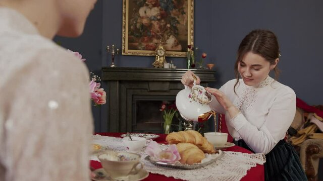 very beautiful girl in a vintage white shirt sits at a beautiful antique table and pours herself tea