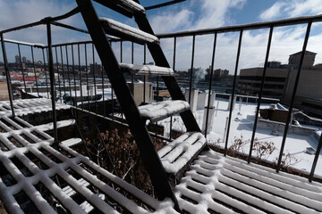 a snow covered fire escape with a cityscape of building tops and a blue cloudy sky in the background
