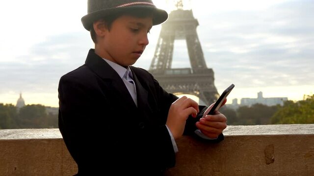 A cute teenage boy in a grey hat is reading a message on the phone screen on the background of the Eiffel tower at the early morning, Paris, France