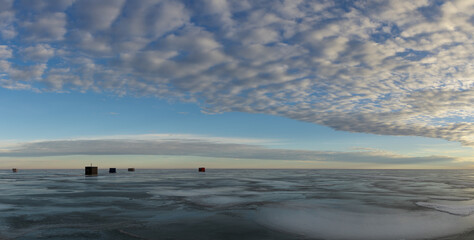Naklejka premium Cold barren looking winter scene with a group of fishing shacks sitting on a large frozen lake with glossy ice and puffy clouds. The ice is reflecting the color of the sky and clouds. 