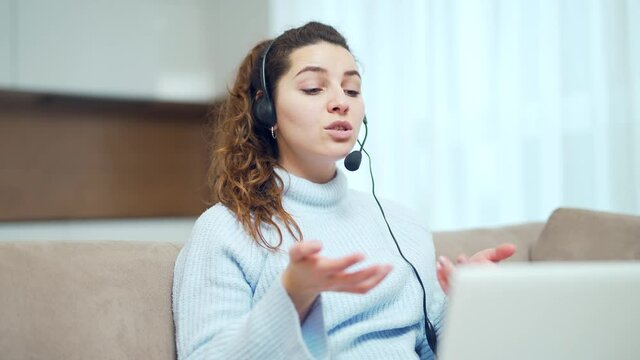 Online distance learning. Woman freelancer at home communicates via internet. The teacher explains remotely. young adult student girl sitting on sofa with headset and laptop and talking to webcam.