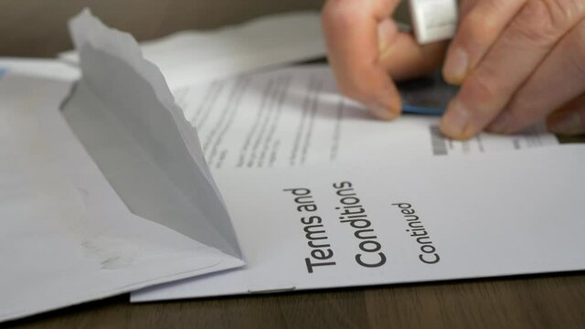 Credit card terms and conditions sharply focused in the foreground, with a defocused background of a man&rsquo;s hands removing a new credit card which is glued to his bank&rsquo;s letter.