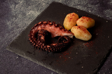 Delicious grilled octopus tentacles with potatoes seasoned with Spanish paprika, olive oil, and sea salt on a black slab and fancy background. Gourmet kitchen concept, culinary aesthetics.