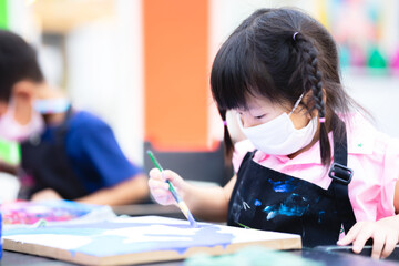 Child wearing cloth mask is in an art class. Girl are working on painting watercolor on the canvas. Children grabs the paintbrush with his right hand. New normal. Prevent spreading the coronavirus.