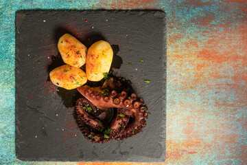 Delicious grilled octopus tentacles with potatoes seasoned with Spanish paprika, olive oil, parsley and sea salt on a black slab and fancy background. Gourmet kitchen concept, culinary aesthetics.