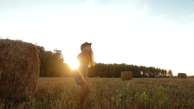 Young girl in denim overalls and a cowboy hat jumps off a bale of straw or hay and poses against the sunset. Child plays in a rural area in Sunny weather in the evening. Slow motion