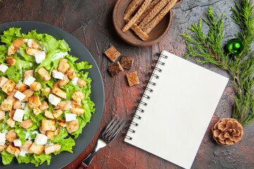 top view caesar salad on oval plate fork bowl with dried crust a notebook on dark red background food photo