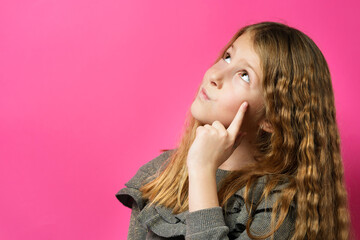 Idea concept - Young thoughtful caucasian girl eight years old looking up with finger on face contemplation in front of pink background - copy space