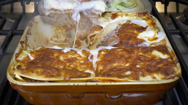 Closeup POV shot of a slice of homemade, freshly cooked, steaming hot lasagne being lifted from a ceramic baking dish on an oven hob, onto a plate of salad behind.