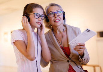 Senior female tutor and tweenage girl exploring art pieces in museum and listening commentaries in...